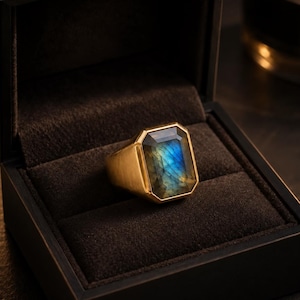 May include: A gold ring featuring a large, rectangular, faceted labradorite gemstone with a prominent blue flash. The ring is displayed inside a dark, velvet-lined box, highlighting the contrast between the metallic gold band and the stone.
