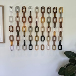May include: A wall display featuring nine vertical chains made of interlocking oval links in various colors, including clear, black, brown, tan, and tortoiseshell patterns. Each chain is suspended from a small, individual wall hook.