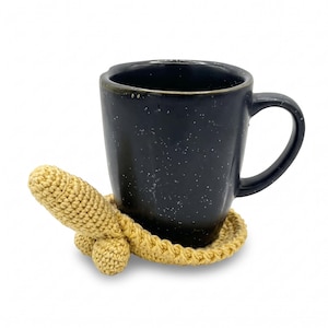 May include: A black ceramic mug with white speckles sits on a beige crocheted coaster. The coaster features a circular base with a three-dimensional, phallic-shaped crochet extension protruding from one side, crafted from light tan yarn.