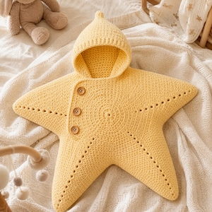 May include: A bright yellow, star-shaped baby swaddle blanket featuring a pointed hood and three brown wooden buttons along the side. The piece is crafted with a textured crochet knit pattern and includes decorative perforated lines along each arm.