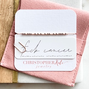 May include: A rose gold beaded bracelet with a message in Morse code. The bracelet is displayed on a white card with the words "fuck cancer" and "CHRISTOPHER Kate jewelry" printed on it. The card is attached to a pink suede pouch.