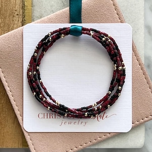 May include: A multi-strand beaded bracelet with red, black, and gold beads. The bracelet is wrapped around a white card with the text "CHRISTOPHER KATE jewelry".