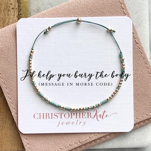 May include: A light blue and gold beaded bracelet with a message in Morse code that reads "I'd help you bury the body". The bracelet is on a white card with the text "CHRISTOPHER Kate jewellery".