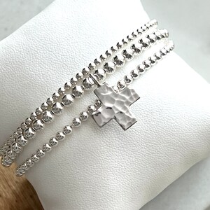 May include: A silver bracelet with three strands of small beads and a hammered silver cross charm.