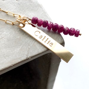 May include: A gold-colored necklace with a rectangular pendant engraved with the name "Callie." The necklace features a chain and a strand of deep red, faceted gemstones. The jewelry is displayed on a gray surface.