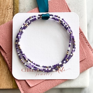 Puede incluir: Un brazalete de cuentas de triple vuelta con cuentas moradas y blancas y detalles dorados. El brazalete se muestra en una tarjeta blanca con el texto "CHRISTINE jewelry".