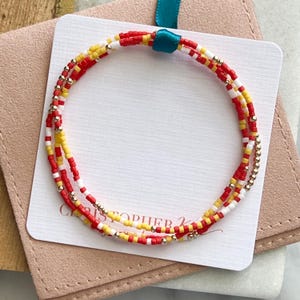 Könnte beinhalten: Ein mehrreihiges Perlenarmband mit roten, gelben und weißen Perlen. Das Armband ist um eine weiße Karte mit dem Text "CHRISTOPHER K" in Schwarz gewickelt.