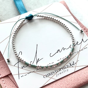 Könnte beinhalten: Silbernes Perlenarmband mit türkisfarbenem Kordel. Das Armband zeigt eine Botschaft in Morsecode und wird auf einer Karte mit den Worten "Fuck Cancer" und "CHRISTOPHER KATE jewelry" präsentiert. Der Hintergrund ist rosa und türkis.