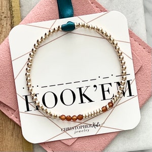 以下が含まれることがあります： 白いカードに「HOOK'EM」の文字と「CHRISTOPHER Kate jewelry」の文字が書かれた、茶色のアクセントビーズが付いたゴールドビーズのブレスレット。