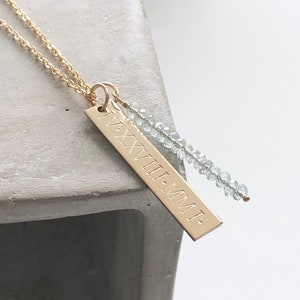 May include: A gold necklace with a rectangular pendant engraved with the Roman numerals "IV.XXVII.MMI." and a strand of light blue beads.