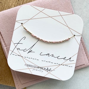 May include: A delicate necklace with pink and gold beads, displayed on a card with the words "fuck cancer" in script. The card is set against a pink background, with the brand name "Christopher Kate Jewelry".