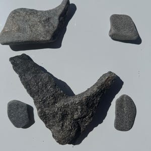 May include: A collection of five grey stones of varying shapes and sizes. One large stone is V-shaped, while the others are smaller, rounded, or elongated. The stones have a rough, textured surface and cast distinct shadows on the surface below.