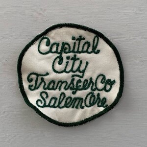 May include: A white, round, embroidered patch with a green border. The text on the patch reads "Capital City Transfer Co. Salem, Ore."