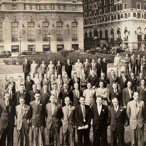 May include: A black and white photograph of a group of men standing in front of a building. The men are all wearing suits and ties. Some of the men are smiling, while others have serious expressions on their faces.