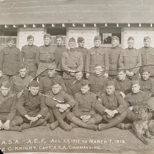 May include: A black and white photograph of a group of men in military uniform, standing and sitting in front of a building. The men are wearing caps and some are holding rifles. The text "A.S.A. - A.E.F., Aug. 23, 1917 to March 7, 1919. E C. KNIGHT, CAPT. A.S.A. COMMANDING." is printed at the bottom of the photo.