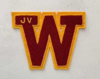 Pode incluir: Uma letra W de feltro vermelho com contorno amarelo. A letra é cercada por uma borda amarela com as letras "JV" em um retângulo branco menor.