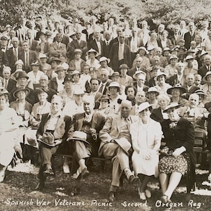 May include: Black and white photo of a group of people, mostly older adults, sitting on a bench outdoors. The group is gathered for a Spanish War Veterans Picnic, Second Oregon Reg. The photo is likely from the early 1900s.