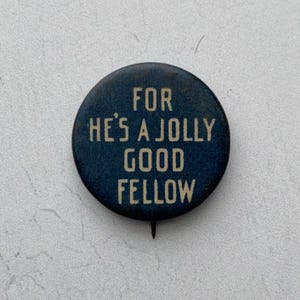 May include: A round, dark blue button with the text "FOR HE'S A JOLLY GOOD FELLOW" in white lettering. The button has a pin on the back and is likely a vintage or novelty item.