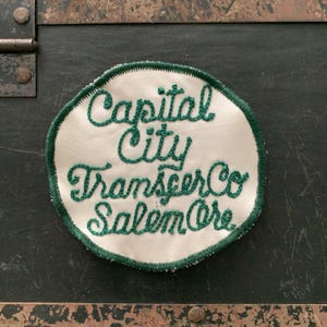 May include: A circular embroidered patch with a cream-colored fabric base and a dark green stitched border. The text Capital City Transfer Co Salem Ore is embroidered in dark green cursive script across the center of the circular patch.