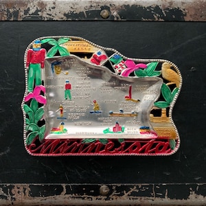 May include: A silver metal souvenir ashtray shaped like the state of Minnesota. The ashtray is decorated with colorful images of landmarks and cities, including International Falls, Grand Marais, Duluth, and St. Paul. The text "Minnesota" is printed in red along the bottom edge.