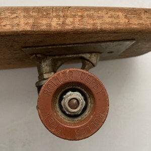 May include: A close-up of a red roller skate wheel with the words "Roller Derby" printed on it. The wheel is attached to a metal axle and a wooden skateboard deck.