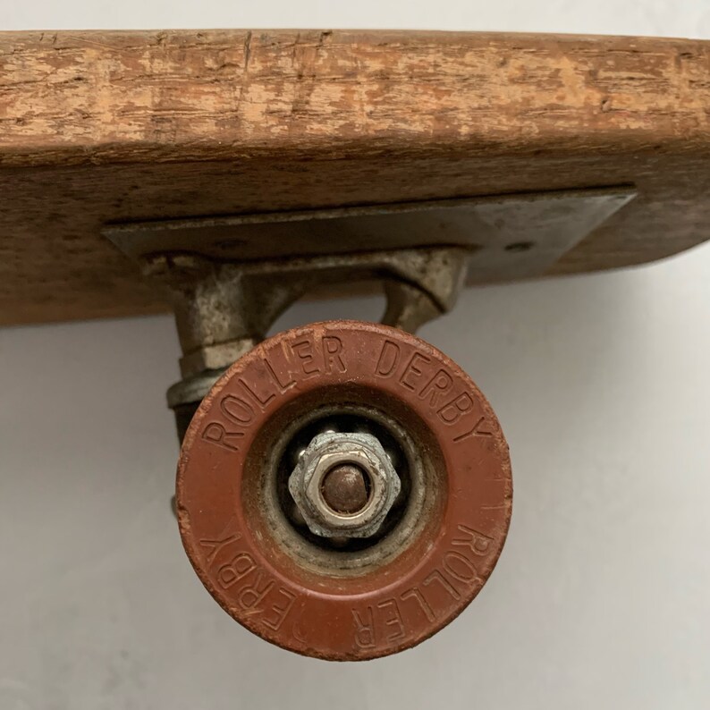 May include: A close-up of a red roller skate wheel with the words "Roller Derby" printed on it. The wheel is attached to a metal axle and a wooden skateboard deck.