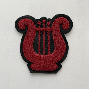 May include: A patch featuring a red, textured, chenille-style lyre musical instrument design centred on a black felt-like backing. The lyre has two curved side arms, a base, and three vertical strings, all rendered in a deep red, fuzzy material.