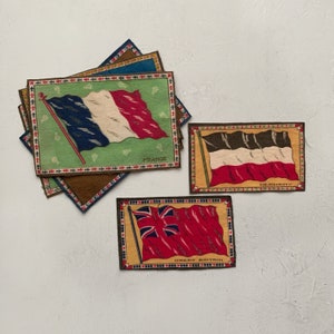 May include: A set of three vintage fabric patches featuring flags of France, Germany, and Great Britain. Each patch is rectangular with a colorful embroidered flag design and a decorative border.
