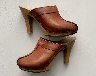 May include: A pair of brown leather heeled clogs with wooden soles. The clogs have a closed toe and a high heel.