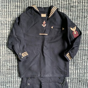 May include: Navy blue sailor suit with a white and tan striped collar and cuffs. The vintage uniform features an embroidered anchor and eagle patch. The jacket has a button-down front and the pants have button details.