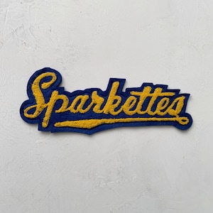 May include: A blue and gold felt patch with the word "Sparkettes" in a script font. The word is in gold, with a blue outline and backing. The patch is likely for a jacket or other apparel.