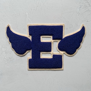 Puede incluir: Parche con la letra "E" azul marino con alas adjuntas. La letra y las alas tienen una apariencia texturizada, con un borde beige claro. El parche es probablemente para una chaqueta o bolso, y está sobre un fondo gris claro.