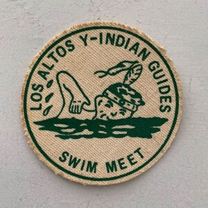 May include: A round, beige fabric patch with green text and a green illustration of a swimmer. The text reads "Los Altos Y-Indian Guides Swim Meet".