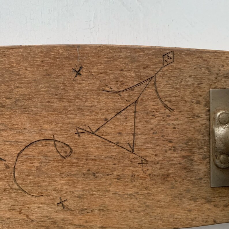 May include: A close-up of a wooden skateboard deck with a triangle and several crosses drawn on it. The deck is brown and has a natural wood grain. The skateboard truck is visible on the right side of the image.