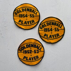 May include: Three vintage yellow and black Goldenball Player patches. Each patch is circular, resembling a basketball, with the text "GOLDENBALL", a year, and "PLAYER" in black lettering. The patches are likely from the 1950s.