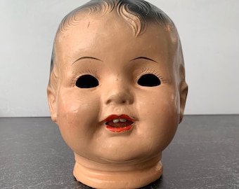 May include: A vintage doll head with a pale peach complexion, molded dark hair, and painted facial features. The doll has hollow black eye sockets, arched eyebrows, long painted eyelashes, and bright red lips with two small white teeth visible in the mouth.