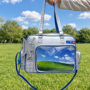 May include: A silver handbag featuring a clear front pocket displaying a green hill and blue sky landscape. The bag includes a silver cursor charm, a side panel with printed computer interface icons, and blue trim. It has two top handles and a shoulder strap.