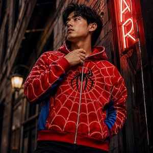 May include: A red zip-up hoodie featuring a black spider emblem on the chest and a white web pattern across the fabric. The garment includes a hood, a full-length silver zipper, and blue side panels, providing a distinct look with long sleeves.