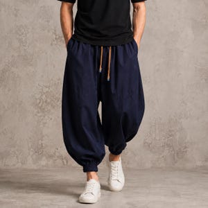 Puede incluir: Pantalones azul marino de corte holgado con cintura elástica alta y cordón marrón. Los pantalones presentan una silueta de pierna ancha que se estrecha en los tobillos con puños elásticos, combinados con una camiseta negra lisa y zapatillas blancas.
