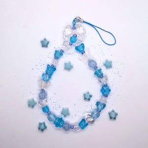May include: A phone charm featuring a loop of blue and clear beads in various shapes, including butterflies and stars. The charm is arranged in a teardrop shape and is surrounded by several small, detached light blue star-shaped beads.