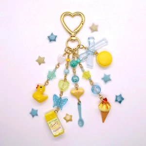 May include: A gold-toned heart-shaped keychain featuring multiple dangling charms including a yellow rubber duck, a miniature milk carton, a blue butterfly, a yellow heart, a small spoon, an ice cream cone, and a yellow macaron with a blue bow.
