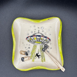 May include: A square ceramic tray with a bright lime green rim and a cream-colored center. The surface features a hand-painted illustration of a flying saucer beaming down a silhouette of a person amidst stars, with two cigarette butts and a joint.