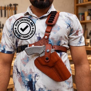 Pode incluir: Um coldre de ombro de couro castanho com uma arma de fogo metálica, apresentando uma alça de ombro com presilhas para munições e uma fivela preta. A camisa exibe um logótipo circular com o texto OUR ORDERS ARE IMPORT TARIFF PAID.