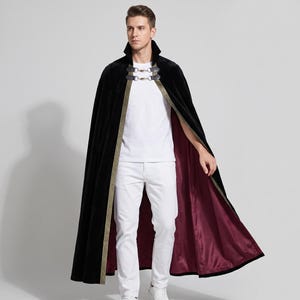 Peut inclure: Une cape longue en velours noir avec une doublure bordeaux profond, un col montant et une fermeture décorative à double agrafe métallique au cou. Les bords avant sont ornés d'une bordure dorée à motif géométrique.
