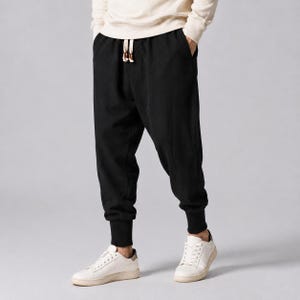 Op de afbeelding: Zwarte joggingbroek met een relaxte pasvorm, voorzien van een tailleband met trekkoord en geribbelde boorden bij de enkels. De broek wordt gecombineerd met een lichtgekleurd shirt met lange mouwen en witte sneakers, inclusief zakken.