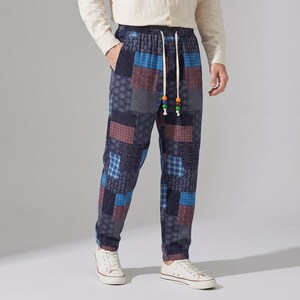 Op de afbeelding: Broek met een patchworkontwerp met diverse blauwe, marineblauwe en bordeauxrode geometrische patronen. De broek heeft een elastische tailleband met een wit trekkoord met kleurrijke kralen aan de uiteinden en een relaxte pasvorm.