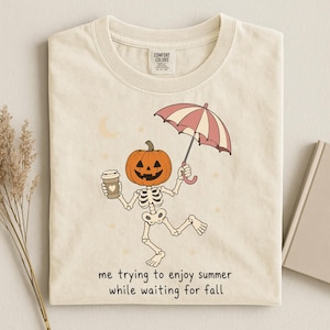 Pode incluir: Uma t-shirt de cor creme com um gráfico de um esqueleto com cabeça de abóbora segurando um copo de café e um guarda-chuva listrado. O texto diz: "me trying to enjoy summer while waiting for fall." A t-shirt tem uma etiqueta Comfort Colors.
