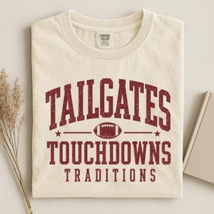 Może przedstawiać: Kremowy t-shirt z krótkim rękawem z bordowym, postarzanym napisem TAILGATES, TOUCHDOWNS oraz TRADITIONS. Grafika przedstawiająca piłkę do futbolu amerykańskiego znajduje się pośrodku, otoczona dwiema małymi gwiazdkami po obu stronach.