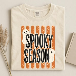 Puede incluir: Una camiseta de color crema con un gráfico de rayas verticales naranjas que contienen puntos blancos. El texto "SPOOKY SEASON" está impreso en letras negras grandes, flanqueado por dos fantasmas de dibujos animados blancos.