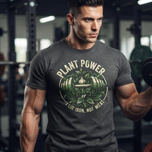 May include: A charcoal gray short-sleeve t-shirt featuring a circular graphic with green leaves surrounding a dumbbell. The text reads PLANT POWER at the top and I LIFT IRON, NOT MEAT at the bottom in a light-colored, bold, sans-serif font.